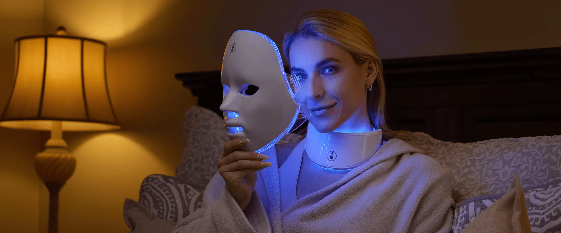A person wears a white RegenaLight LED mask and a matching neck device, both illuminated with vibrant red and blue therapeutic lights against a dark background. The individual's hands are positioned on the sides of the facial mask, highlighting its modern design.